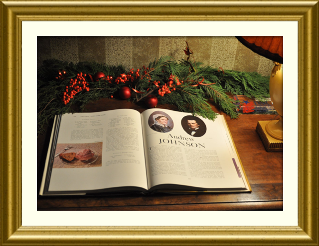 A book about the Johnson family lies open under a lamp. A holly bough is behind the book on the desk.